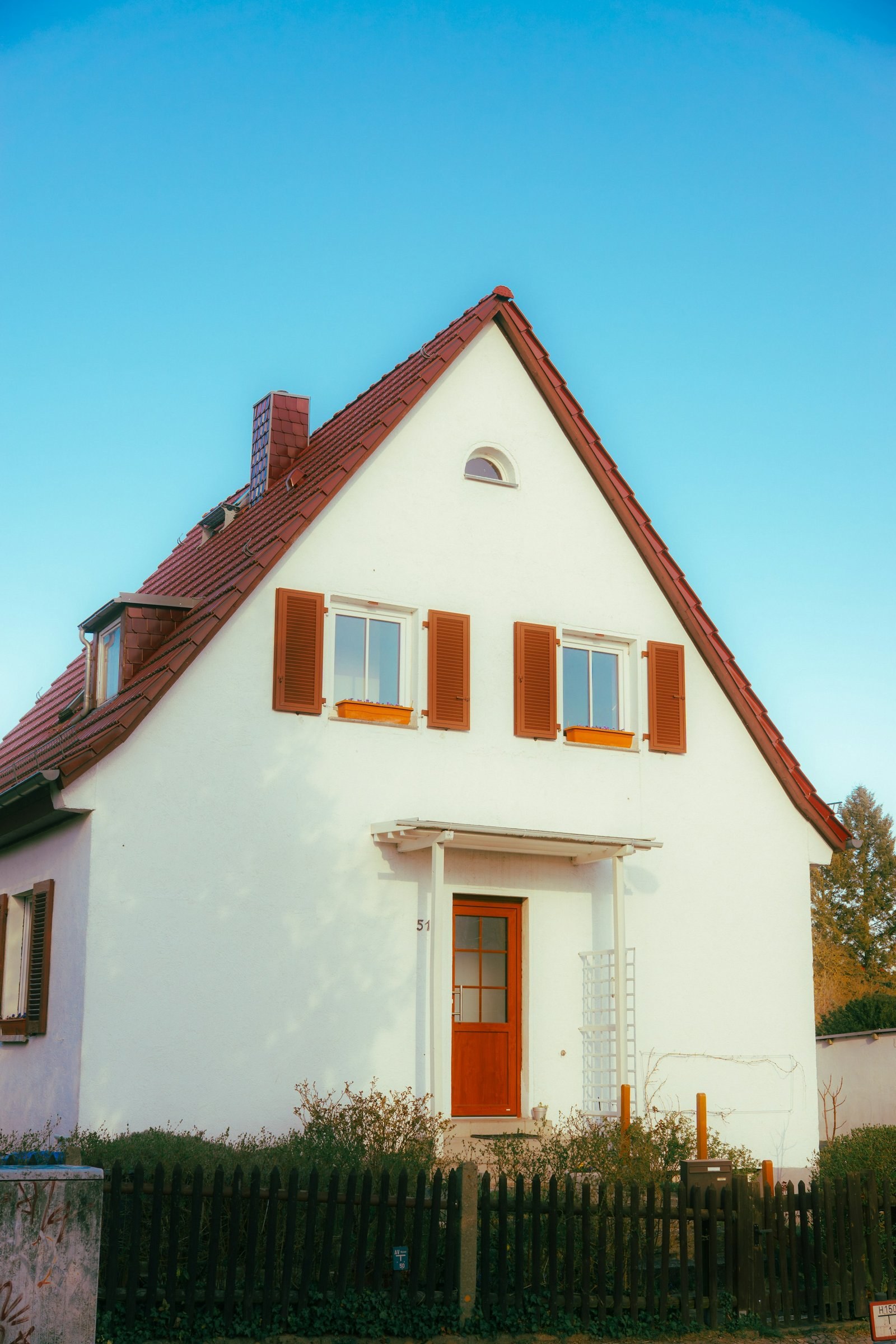 Weißes Einfamilienhaus mit steilem Satteldach aus roten Ziegeln, braunen Fensterläden und Holzzaun vor blauem Himmel