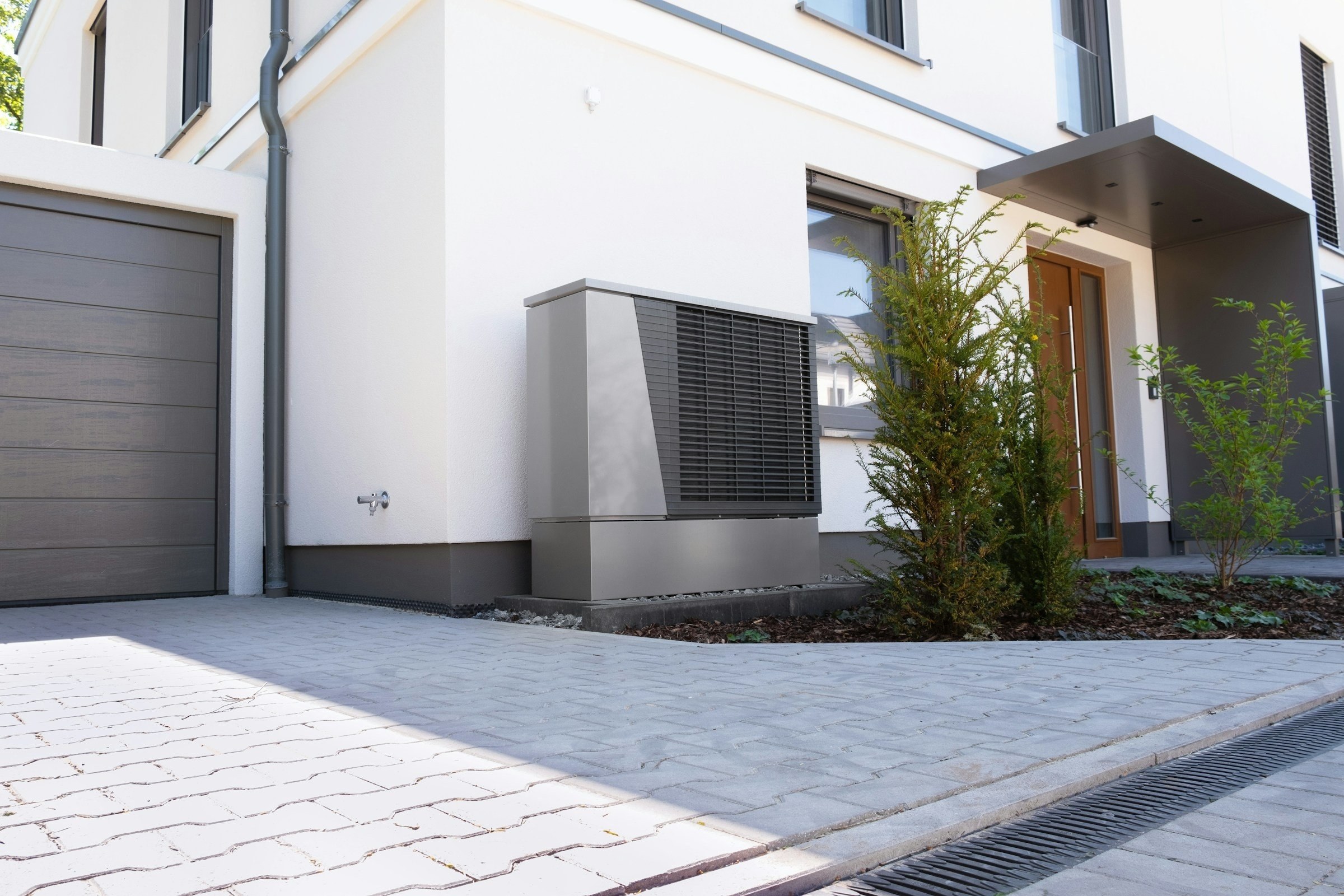Graues Luft-Wasser-Wärmepumpen-Außengerät vor der weiß verputzten Fassade eines modernen Einfamilienhauses, daneben Garagentor, gepflasterte Einfahrt und junge Sträucher