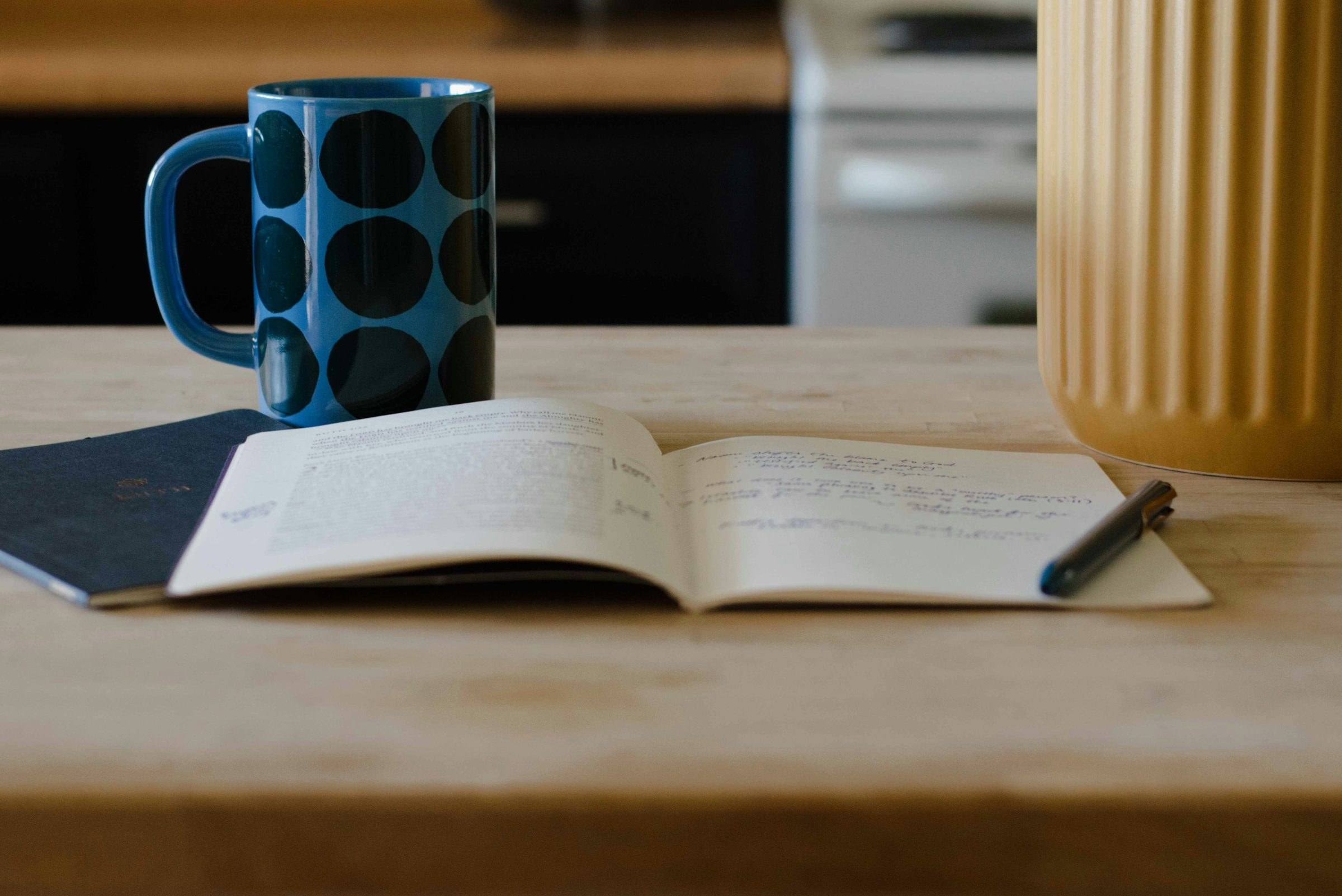 Blaue Tasse mit dunklen Punkten neben einem aufgeschlagenen Notizheft mit handschriftlichen Notizen und einem Stift auf einem hellen Holztisch in einer Küche