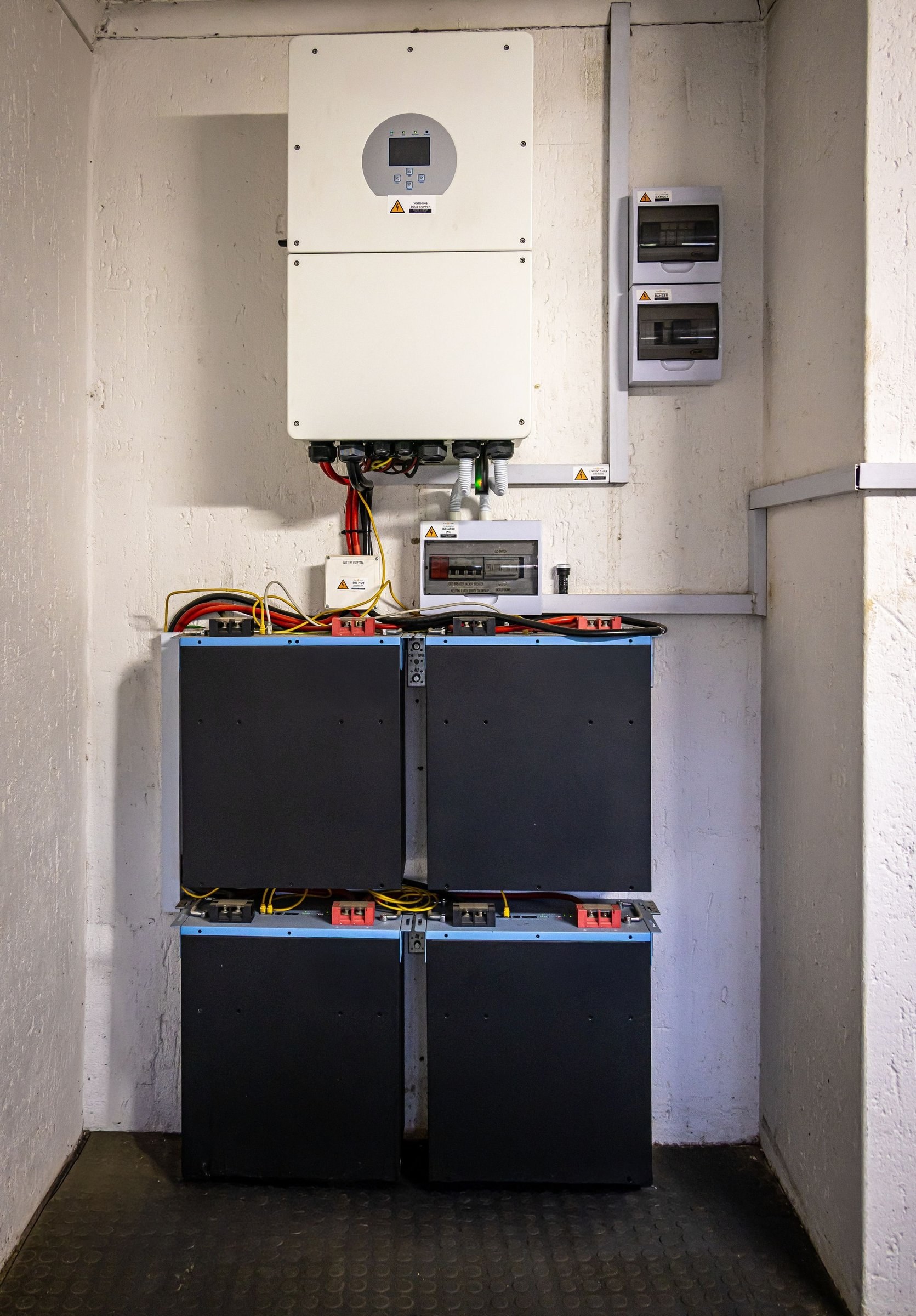 Technikraum mit weißem Wechselrichter an der Wand, Sicherungskästen und vier gestapelten Batteriespeicher-Modulen auf dem Boden
