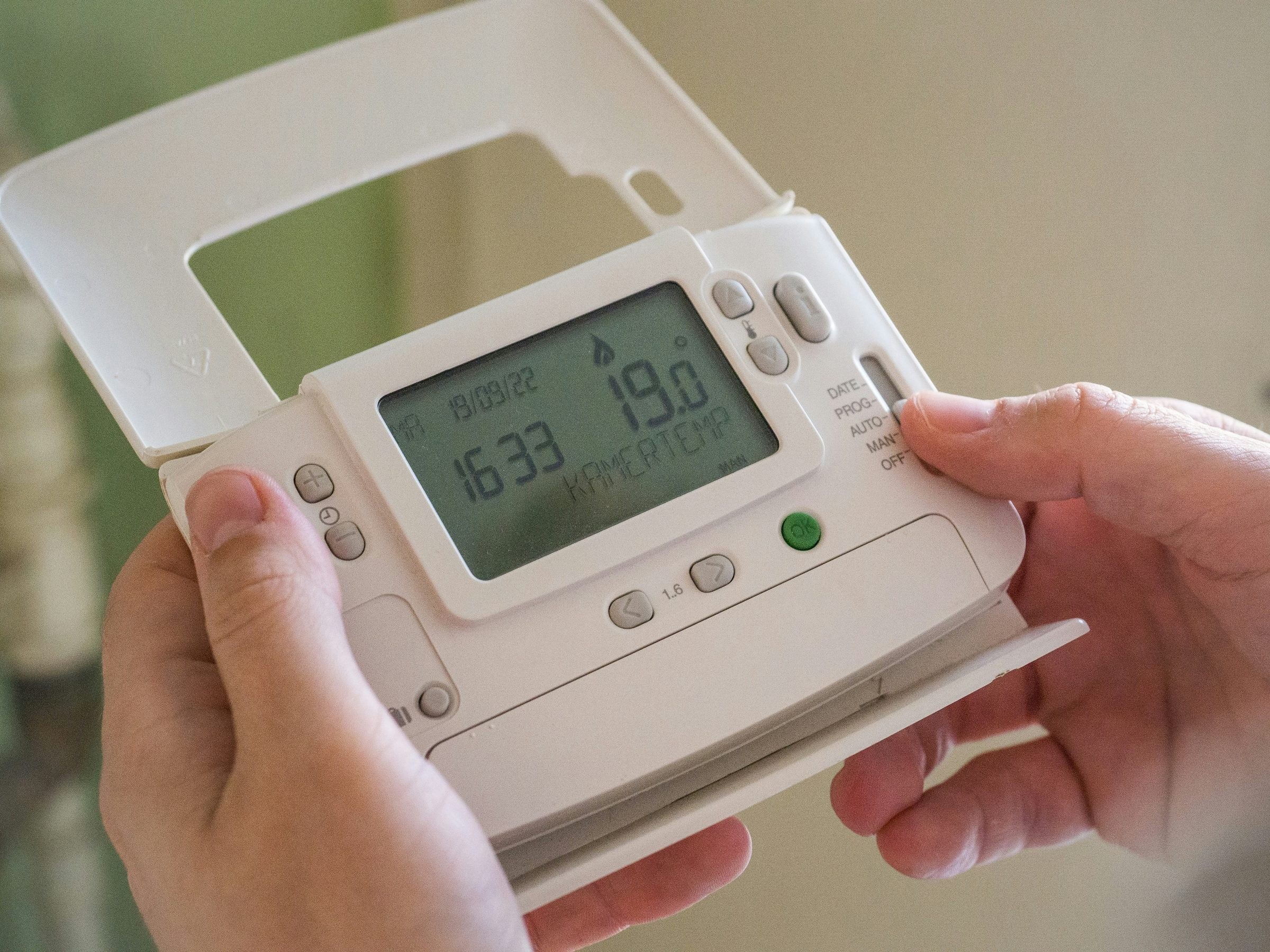 Zwei Hände halten ein aufgeklapptes digitales Raumthermostat mit Anzeige 19,0 Grad und stellen am Schiebeschalter die Betriebsart ein