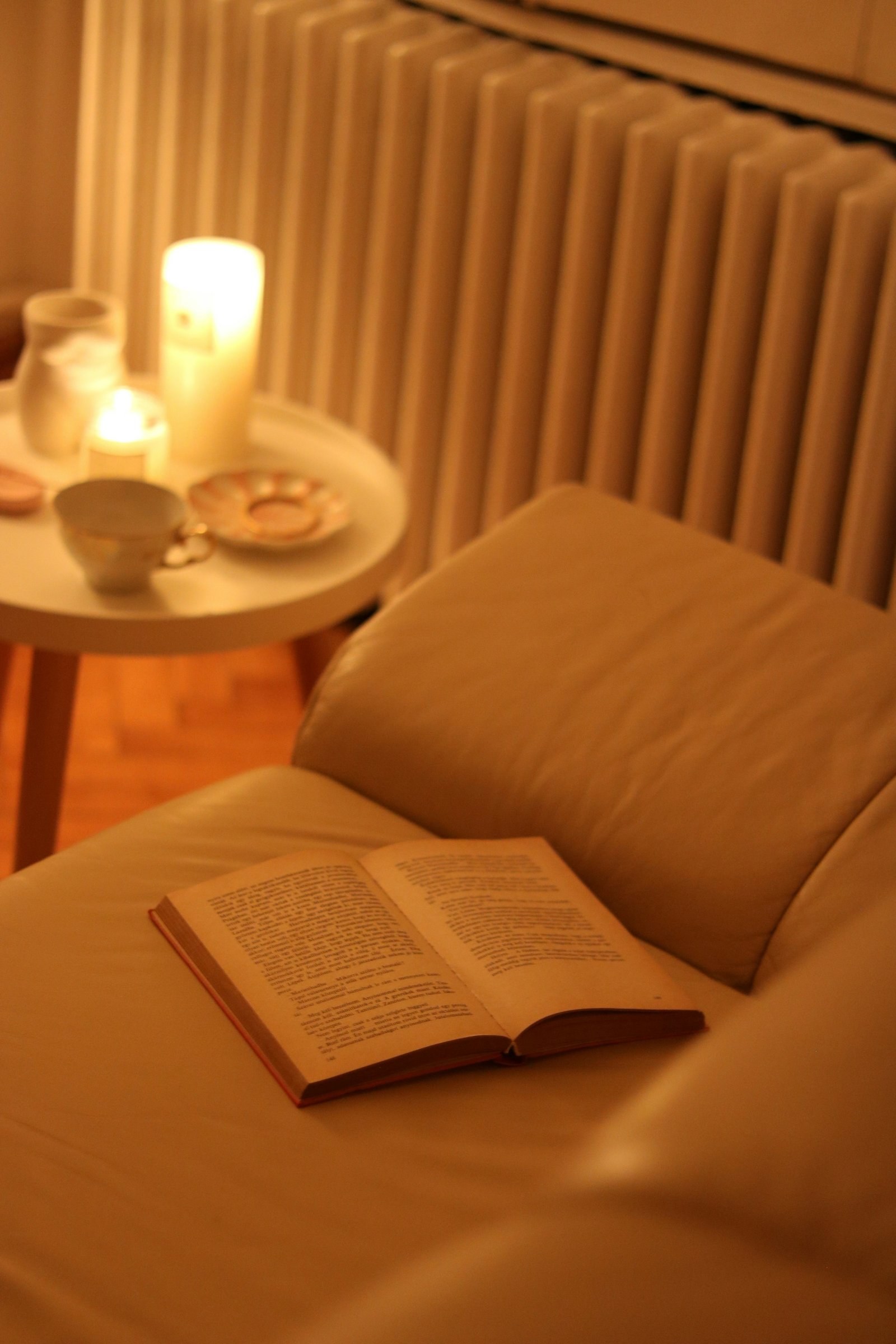 Aufgeschlagenes Buch auf einem Ledersessel, dahinter ein Beistelltisch mit Kerzen und ein weißer Gliederheizkörper im warmen Abendlicht