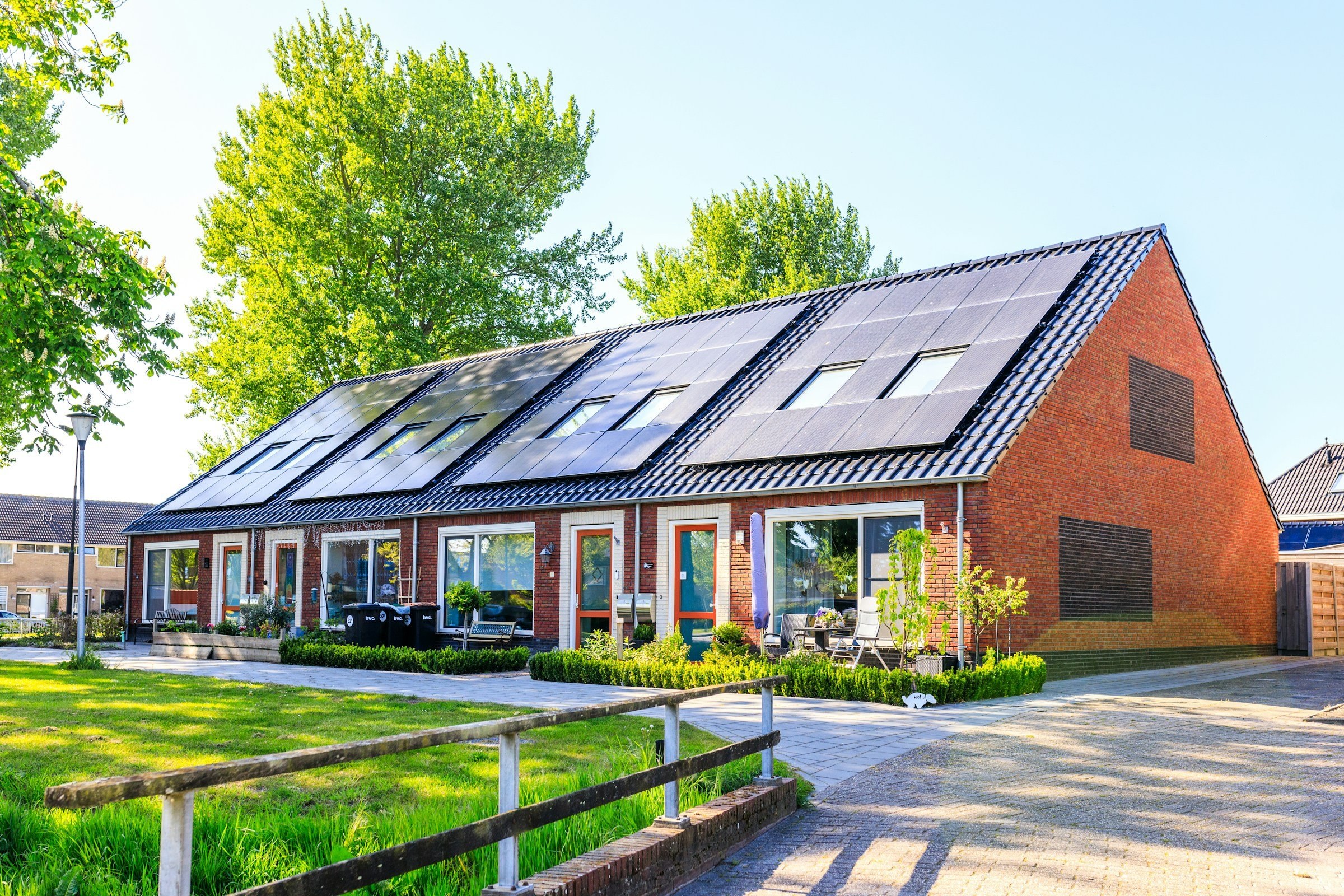 Reihenhauszeile aus rotem Klinker mit dunklen Photovoltaikmodulen und Dachfenstern auf dem Satteldach, davor Rasen und Vorgärten im Sonnenlicht