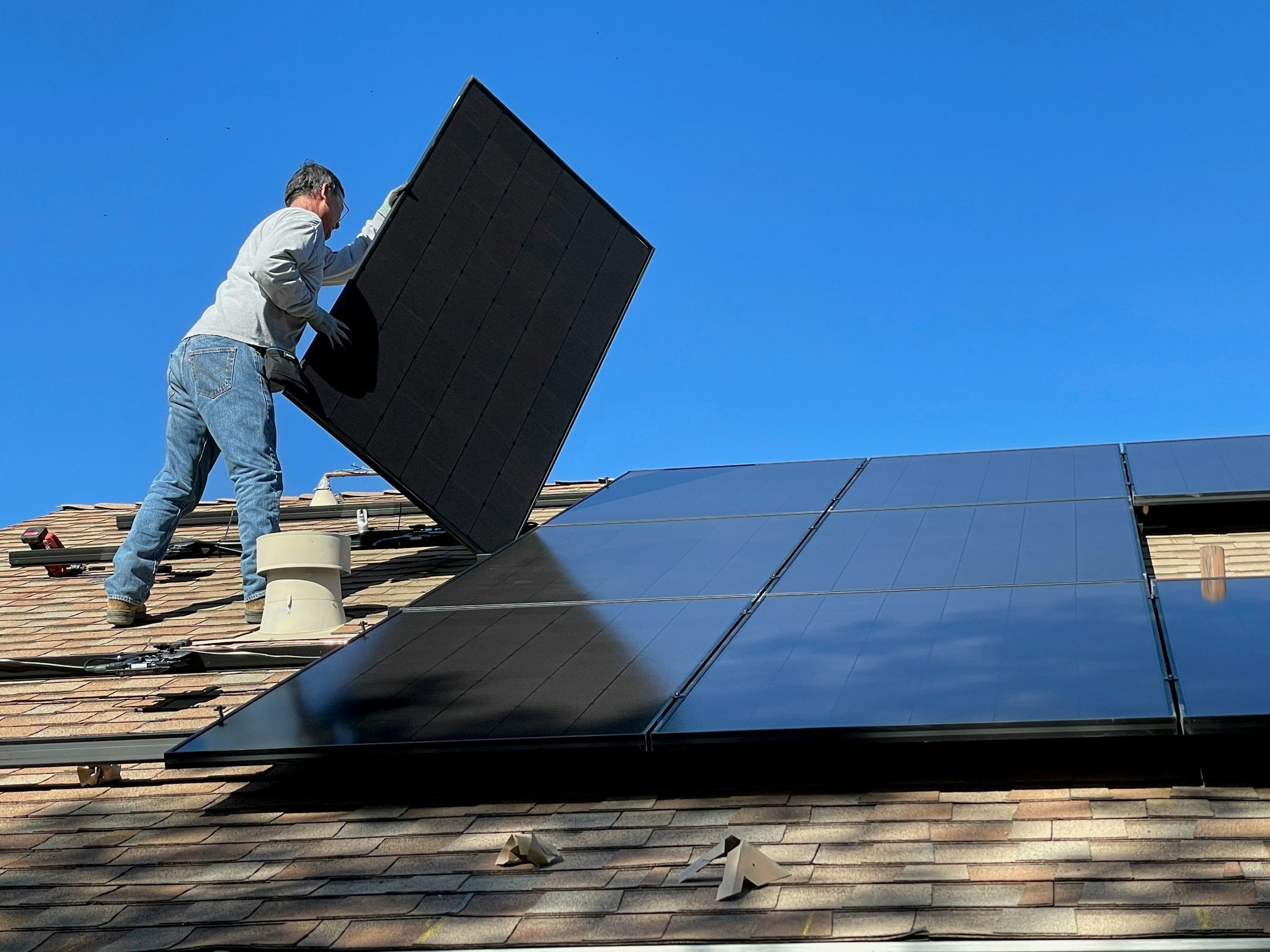 Handwerker auf einem Schrägdach setzt ein schwarzes Solarmodul neben bereits montierte Module, dahinter blauer Himmel